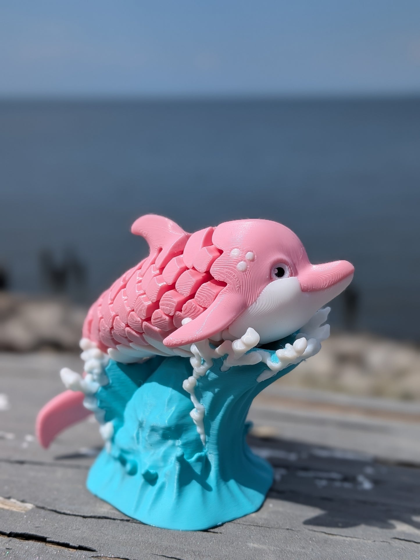 A pink and white dolphin figurine with a blue wave base, displayed outdoors on a wooden surface with a blurred ocean background.