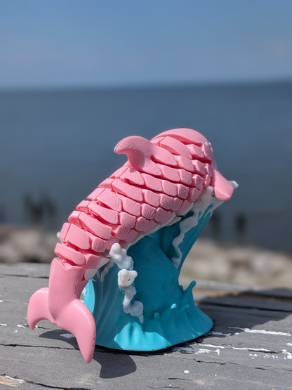 A pink mermaid-themed dolphin sculpture with a blue and white wave base, photographed outdoors on a wooden surface with a blurred ocean background.