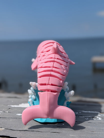 A pink mermaid tail toy or decorative figure with a segmented fin pattern, standing upright on a blue base with white wave details, photographed outdoors against a blurred ocean and sky background.