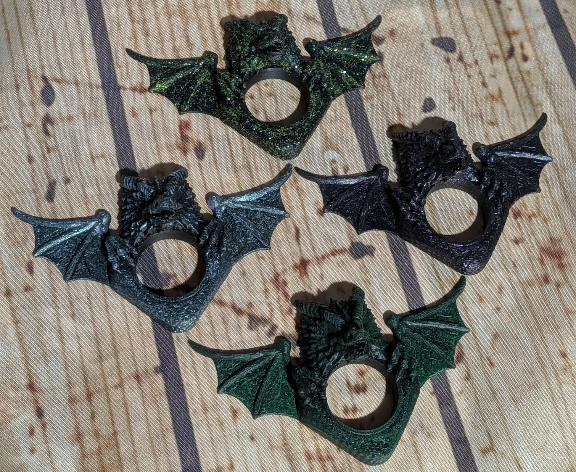 Four bat-shaped ring holders with wings spread, shown in dark metallic and textured finishes on a wood-grain background.