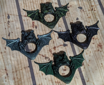 Four bat-shaped ring holders with wings spread, shown in dark metallic and textured finishes on a wood-grain background.