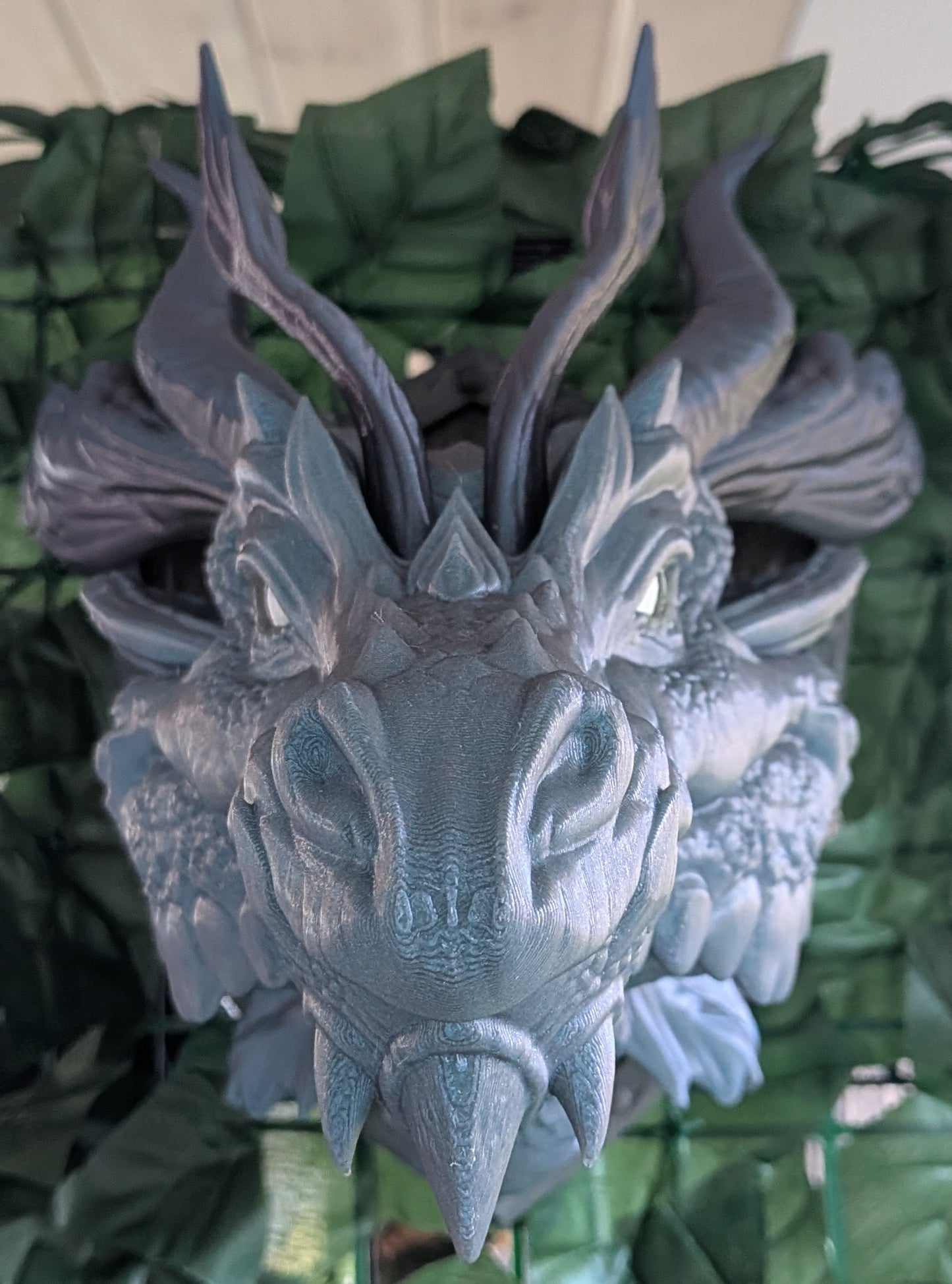 A gray 3D-printed dragon head wall sculpture with detailed scales, horns, and glowing green eyes, set against a background of green artificial leaves.