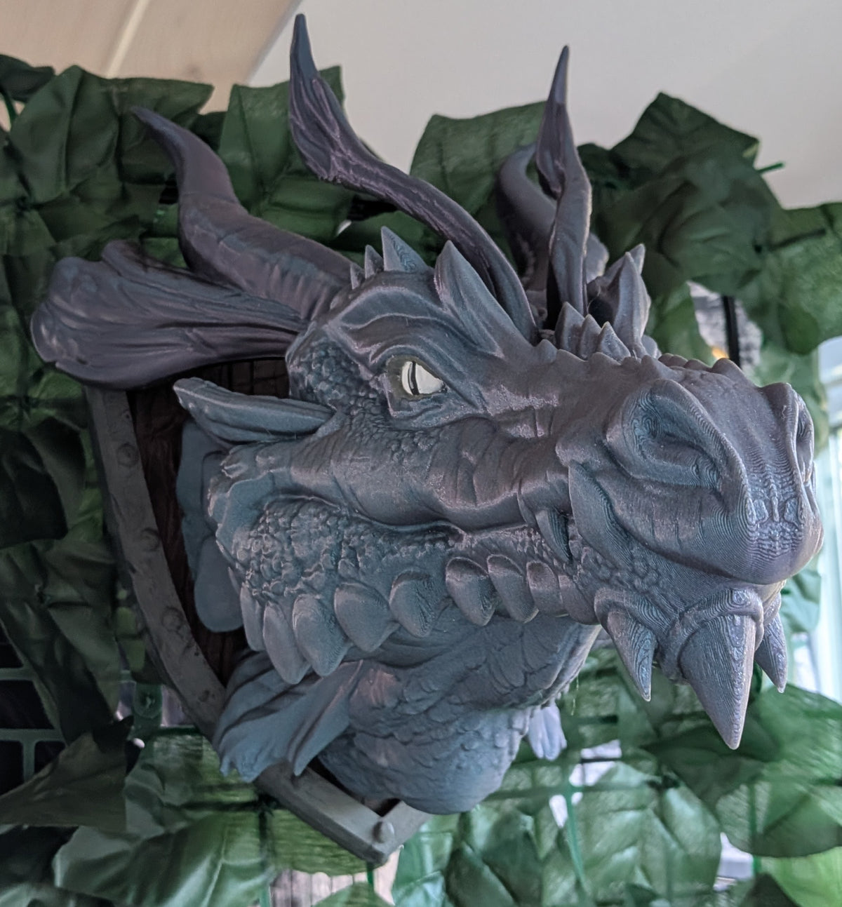 A detailed 3D-printed dragon head wall mount sculpture, shown in a close-up view with green artificial foliage in the background.