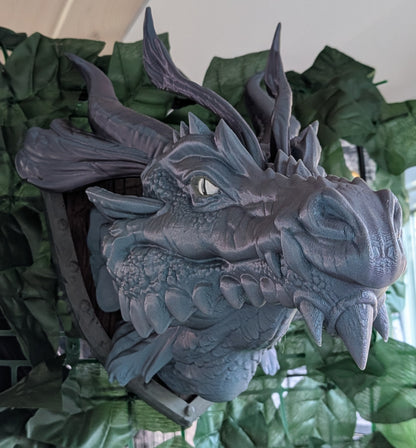 A detailed 3D-printed dragon head wall mount sculpture, shown in a close-up view with green artificial foliage in the background.