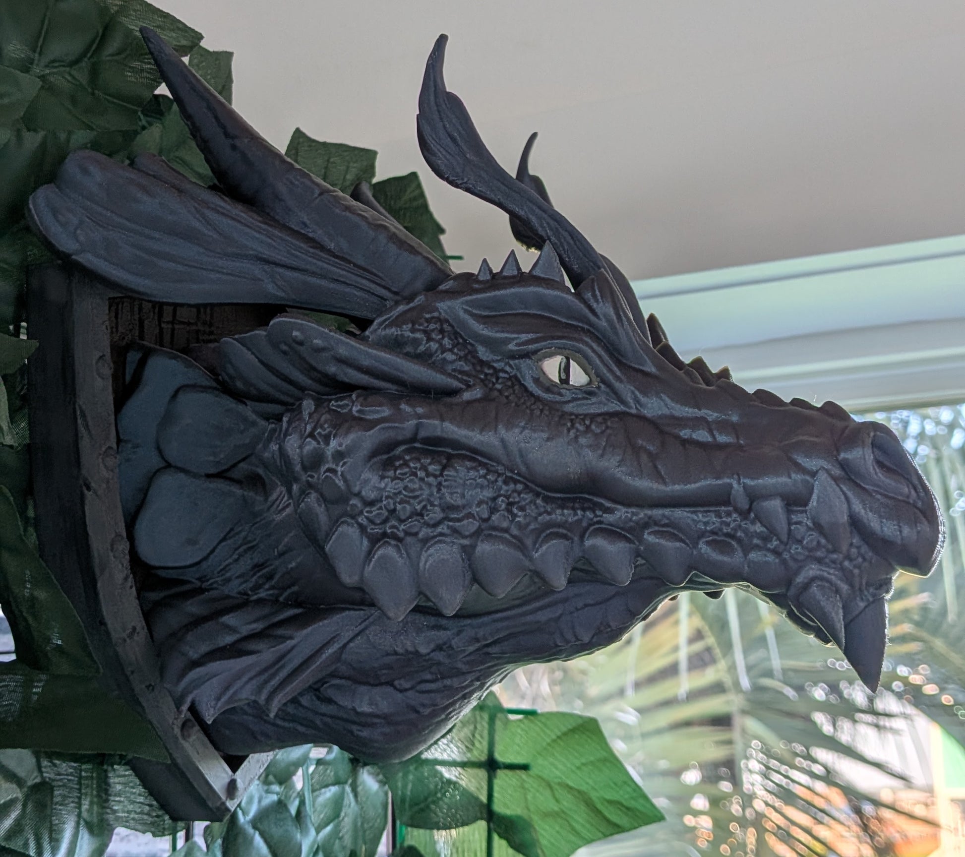 A large dark gray dragon head wall sculpture mounted on a green leafy display, photographed indoors near a window.