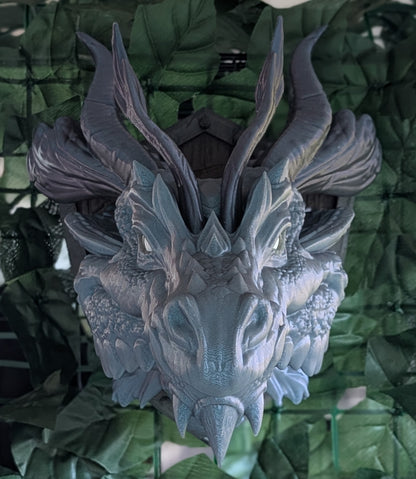 A detailed fantasy dragon head wall hanging or decorative sculpture, featuring textured scales, prominent curved horns, and a weathered gray-blue finish, set against a background of green artificial leaves.