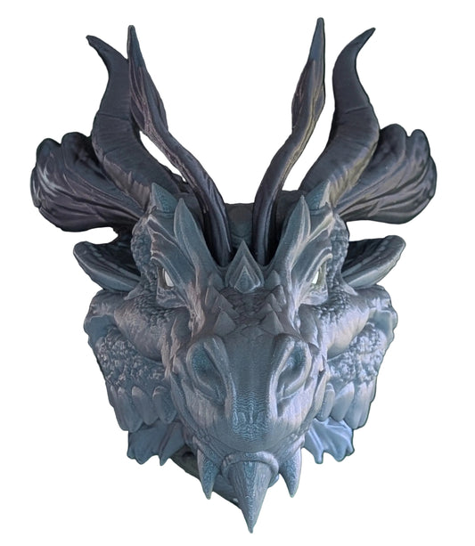 A detailed fantasy dragon head wall hanging or decorative sculpture, featuring textured scales, prominent curved horns, and a weathered gray-blue finish, set against a background of green artificial leaves.