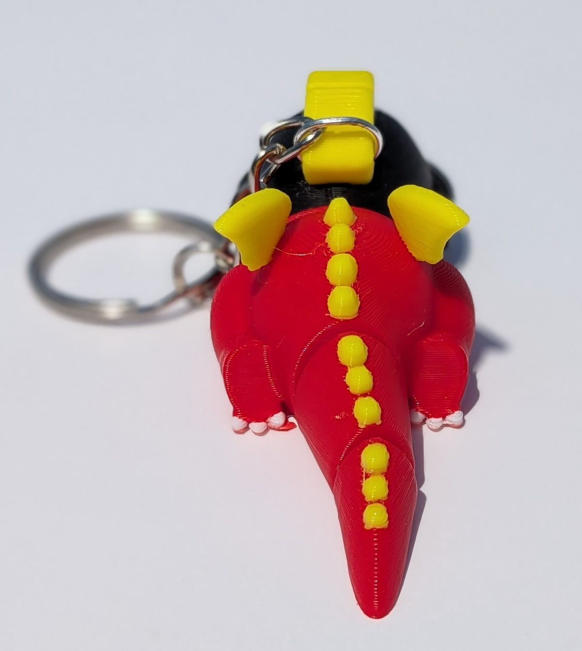 A 3D-printed dinosaur keychain shaped like a red sauropod with bright yellow spikes and horns, attached to a silver metal keyring on a plain white background.