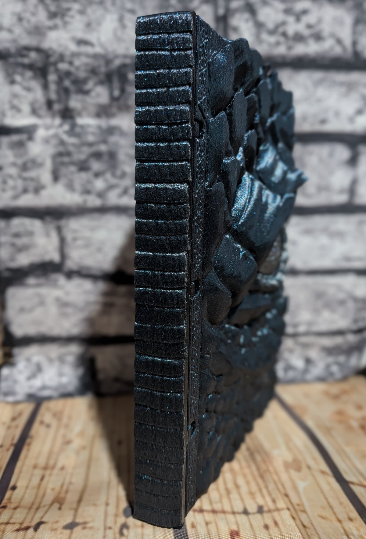 A black cast iron decorative wall plaque featuring a detailed relief carving of a dragon, shown standing upright on a wooden surface against a gray brick wall background.