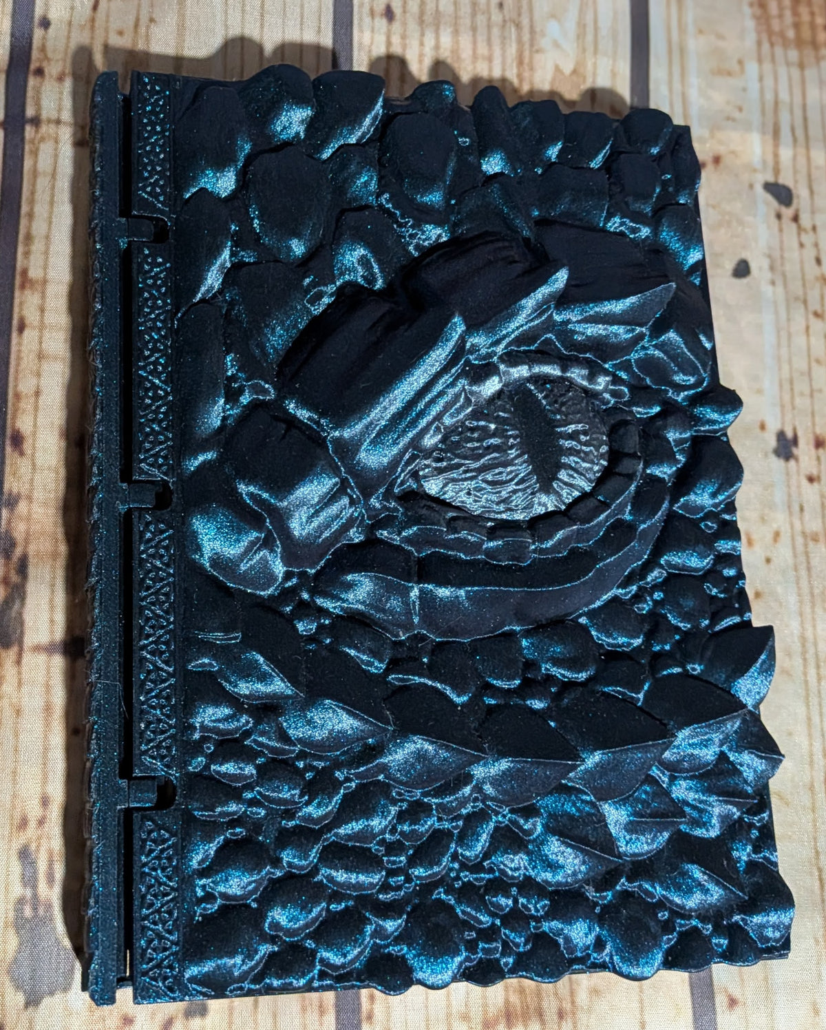 A black textured hardcover journal with a raised, sculpted eye motif on the cover, photographed from above on a light wood surface.