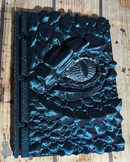 A black textured hardcover journal with a raised, sculpted eye motif on the cover, photographed from above on a light wood surface.
