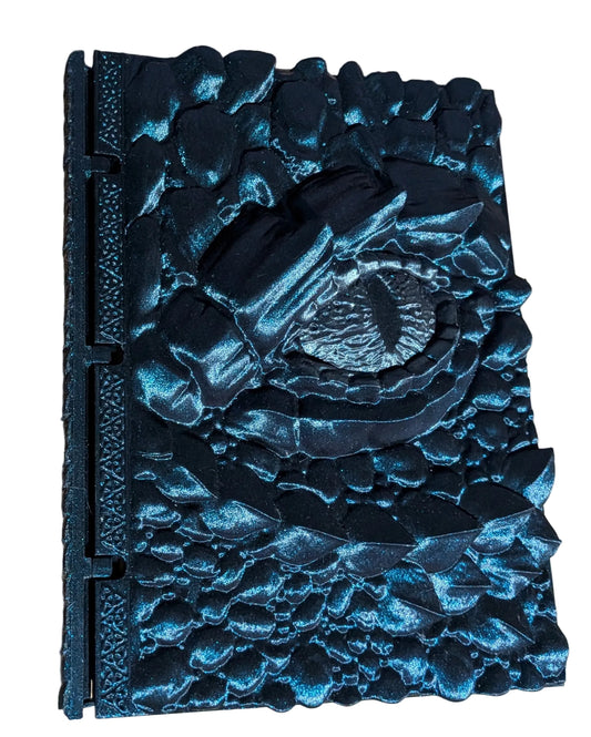 A black textured hardcover journal with a raised, sculpted eye motif on the cover, photographed from above on a light wood surface.