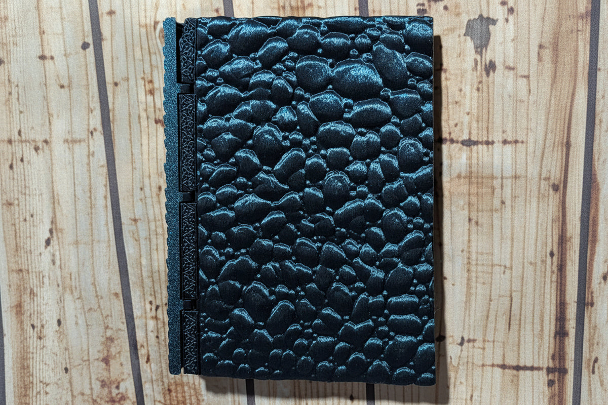 A dark teal textured journal with a pebble-embossed cover and a matching spine, shown on a light wood background.