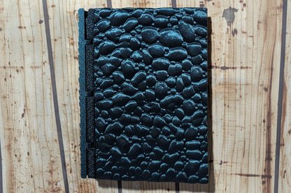 A dark teal textured journal with a pebble-embossed cover and a matching spine, shown on a light wood background.
