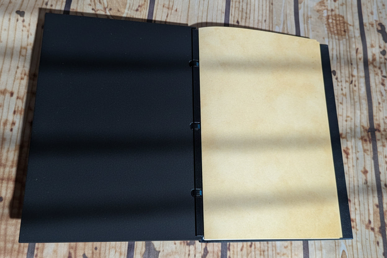 An open handmade ring-bundled journal with a black cloth cover and blank beige paper pages, displayed on a rustic wooden surface.