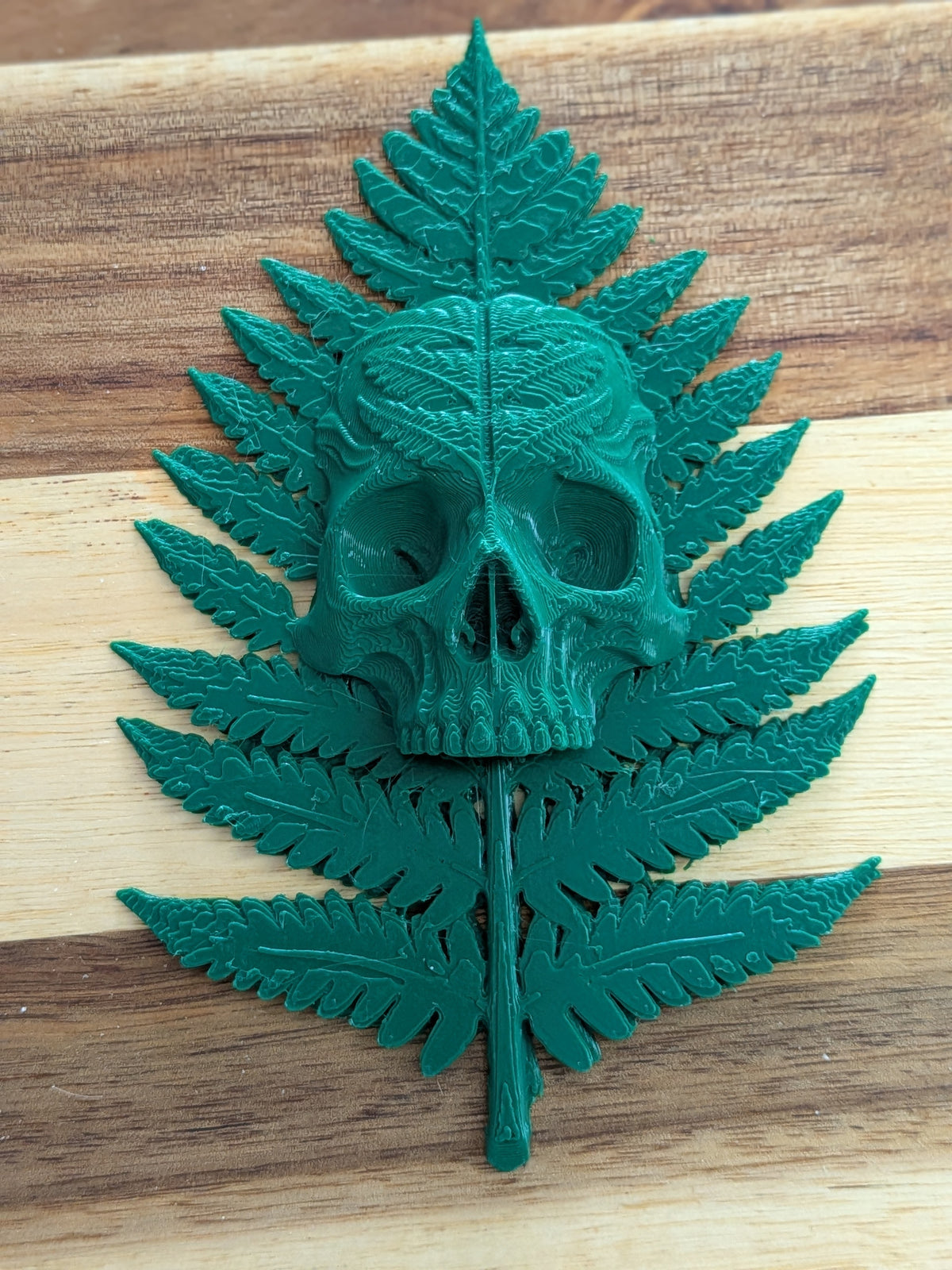 A dark green skull and fern leaf design, likely a 3D-printed patch or pin, displayed on a wooden surface.