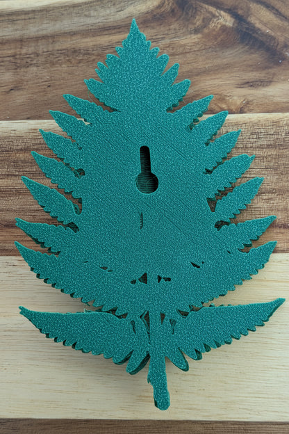 A green fern-shaped decorative object with a keyhole cutout in the center, placed on a wooden surface. A small white price tag reading $5 is attached near the middle.