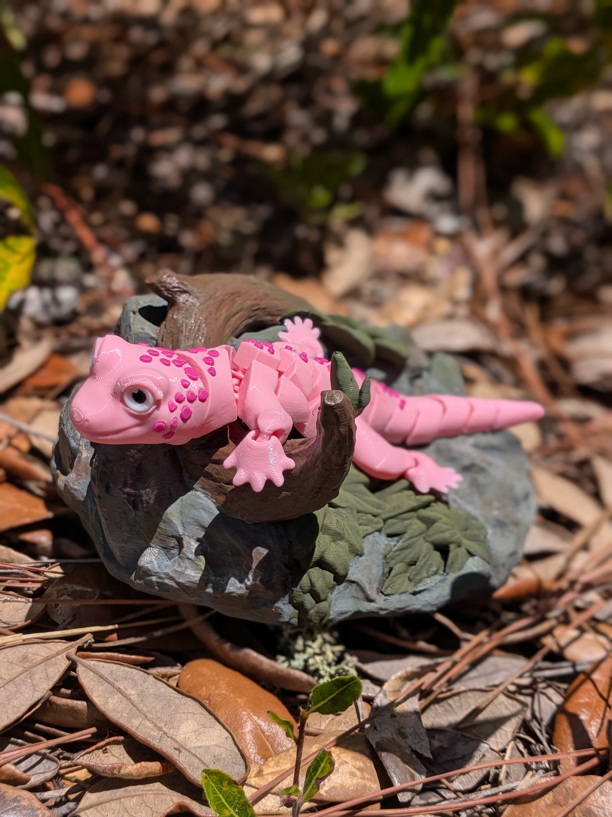 A pink toy gecko figurine with textured spots, posed on a small dark brown rock with green moss, set on a bed of dry leaves and pine needles outdoors.