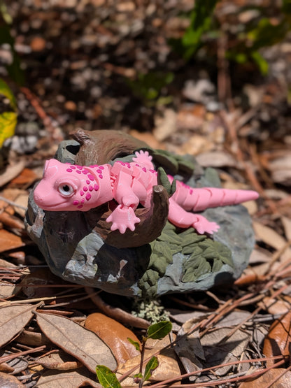 A pink toy gecko figurine with textured spots, posed on a small dark brown rock with green moss, set on a bed of dry leaves and pine needles outdoors.