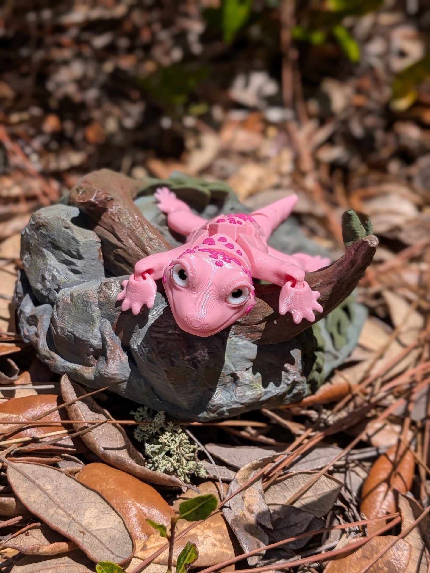 A small pink gecko figurine with darker pink spots, posed on a gray rock sculpture outdoors on a bed of dry brown leaves and pine needles.