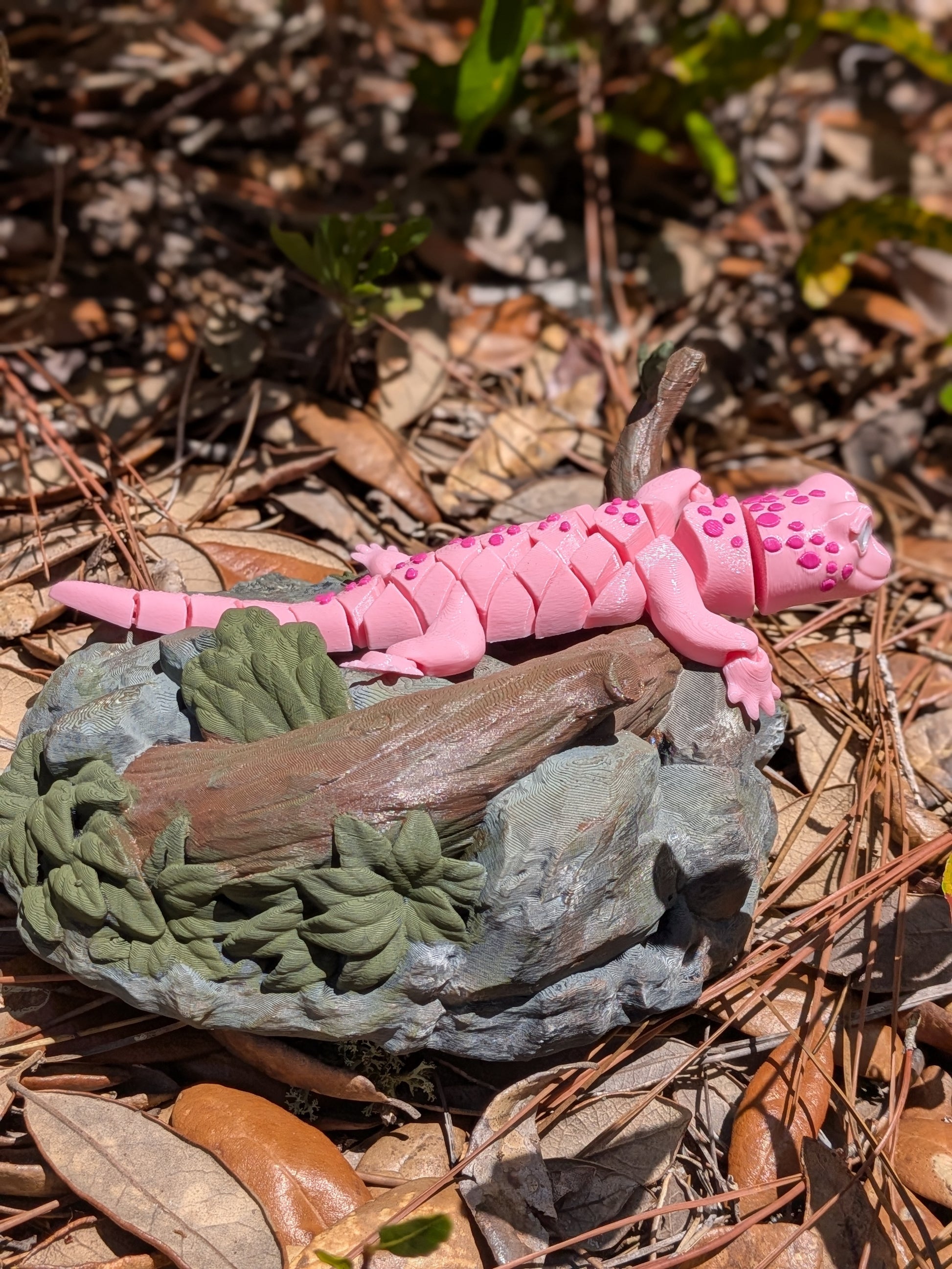 A pink segmented toy salamander figurine resting on a small gray rock with faux moss and a branch, placed outdoors on a bed of pine needles and dry leaves.