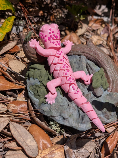 A pink decorative lizard figurine with raised scale details, posed on a gray rock with green leaves and dried pine needles in an outdoor natural setting.