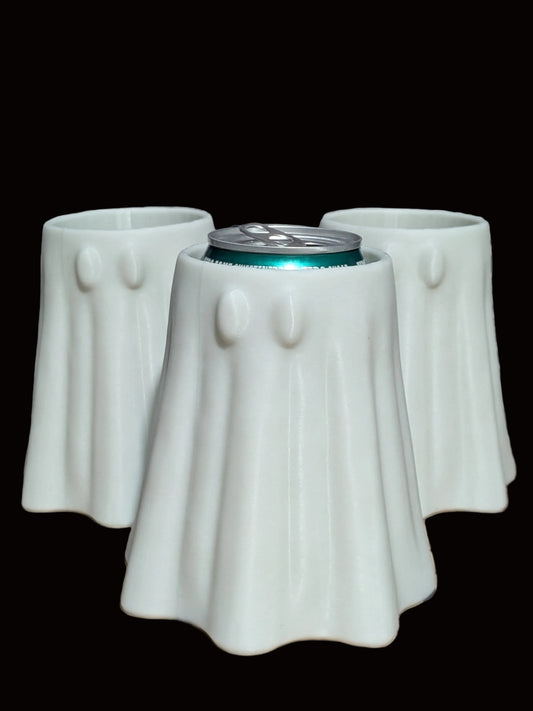 Three ghost-shaped ceramic can coolers in a light mint green color, arranged on a rustic wooden surface with a gray brick wall background. The center cooler is shown holding a silver can with a teal glow emanating from inside.
