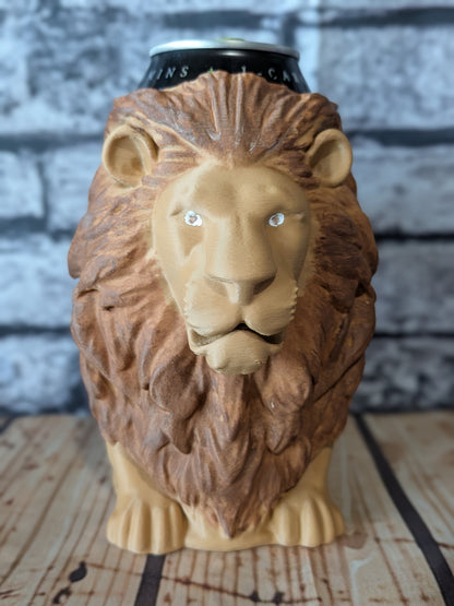 A lion-shaped ceramic mug or can holder with a textured brown mane and tan face, shown holding a black can at the top, set on a wooden surface against a gray brick wall background.