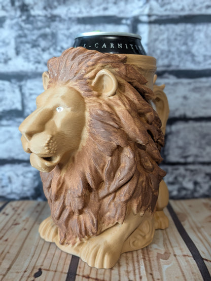 A lion-head mug shaped like a lion, holding a black can with white lettering, set on a wooden surface against a gray brick wall background.