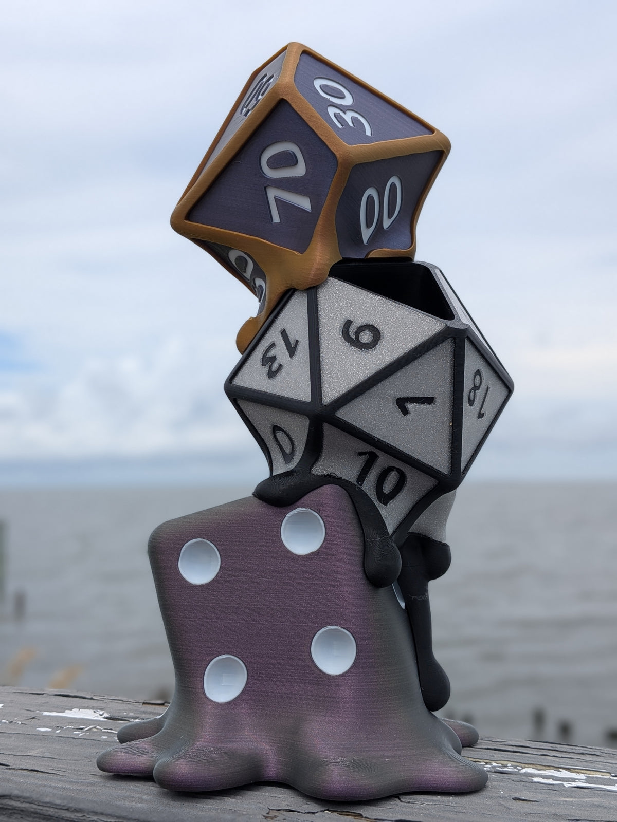 Melting dice planter: a giant D100, D20, and D6 dripping down a rocky drip-tray base, front view