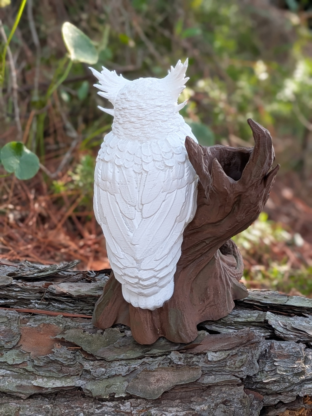 A white painted owl figurine perched on a brown tree stump base, photographed outdoors on a log with blurred greenery in the background.