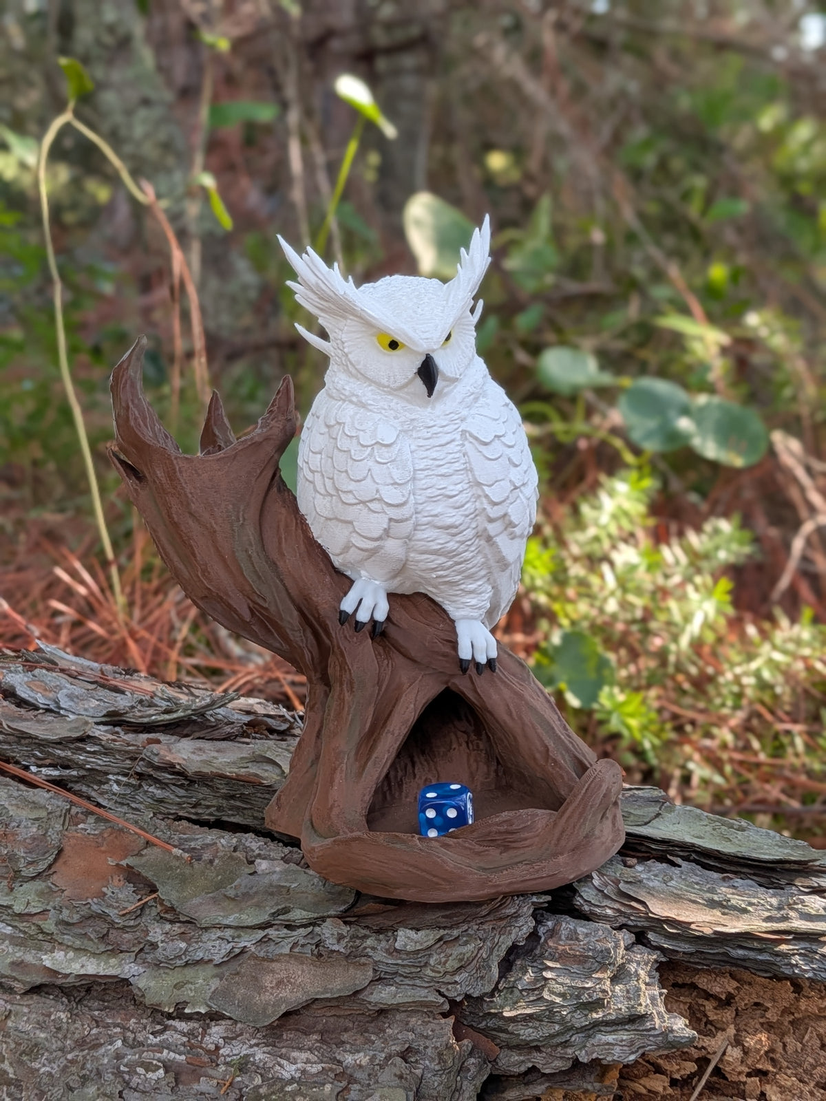 A white owl figurine with yellow eyes and a black beak perched on a brown hollowed tree stump, with a small blue die inside the stump, set outdoors on a log with blurred green foliage in the background.