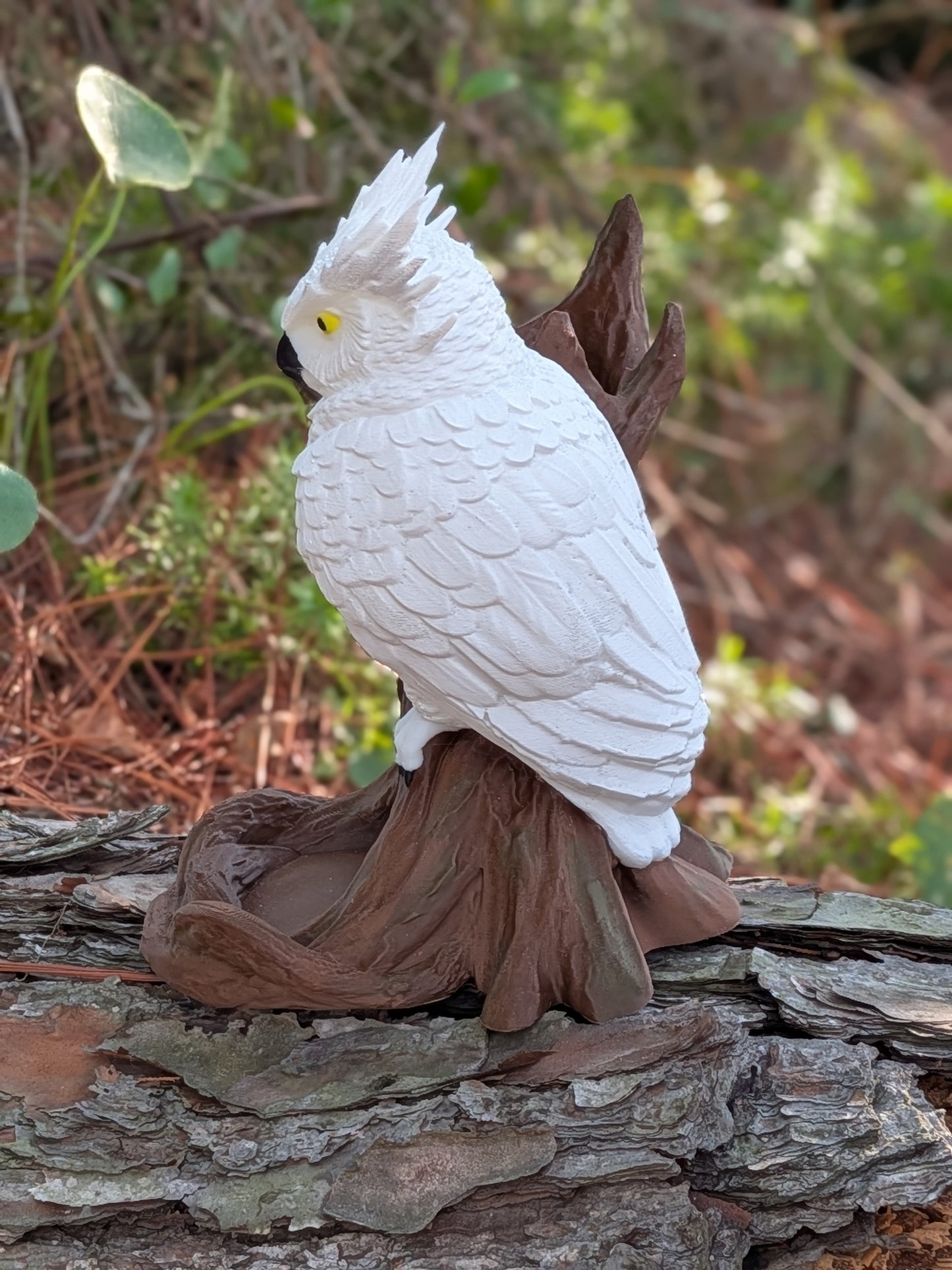 A white cockatoo figurine perched on a brown tree stump base, photographed outdoors against blurred greenery and wood.