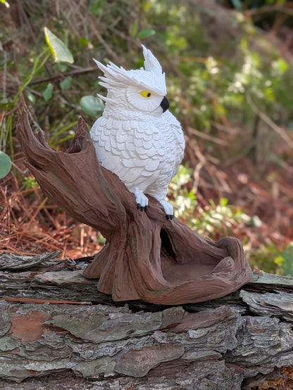 A white cockatoo garden statue perched on a brown faux tree trunk, set outdoors on a stone surface with a blurred natural background.