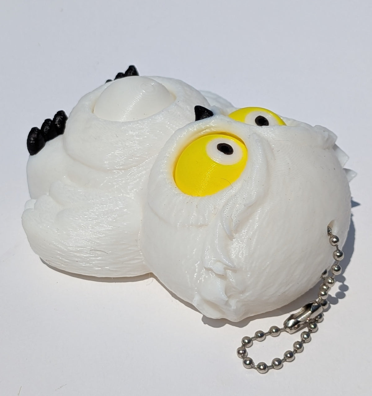 A white, textured owl-shaped squishy toy with large yellow eyes, small black accents, and a silver ball chain, photographed on a plain white background.