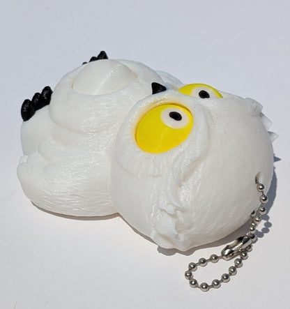 A white, textured owl-shaped squishy toy with large yellow eyes, small black accents, and a silver ball chain, photographed on a plain white background.
