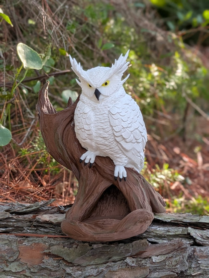 A white owl figurine with yellow eyes and black beak details, perched on a brown textured branch sculpture, set outdoors on a log with blurred green foliage in the background.