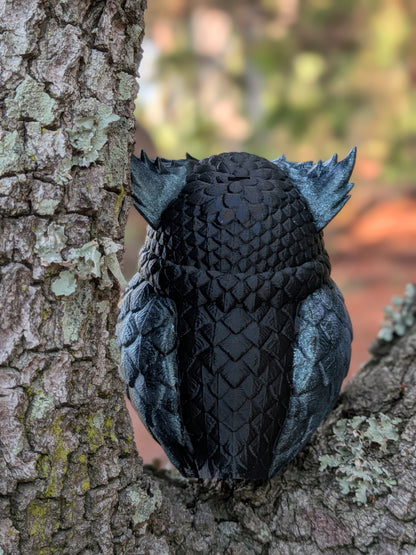 A black owl figurine with textured, scale-like wings and ear tufts, perched on a tree branch next to a tree trunk in an outdoor setting.