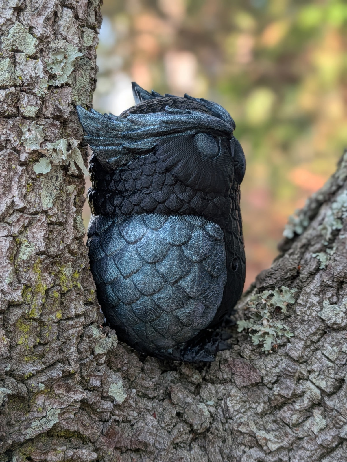 A realistic black and gray kookaburra toy or figurine nestled in a tree branch opening, with textured bark on the left and a blurred outdoor background.