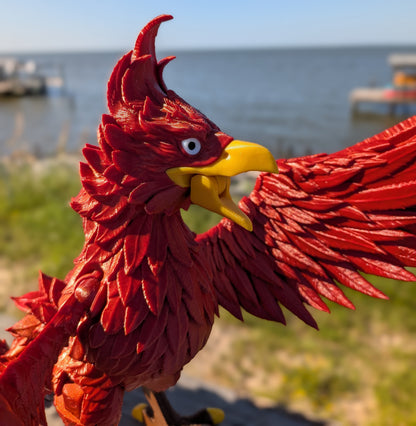 A close-up of a red phoenix bird figurine with a yellow beak and white eye, set against a blurred outdoor background of water, grass, and sky.