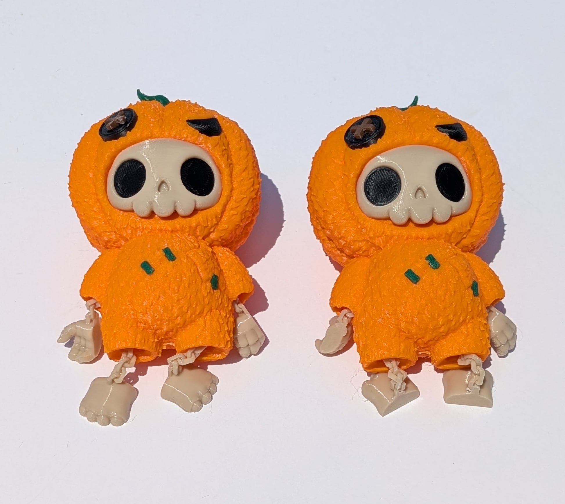 Two small orange pumpkin-shaped figurines with skull faces, black eyes, and beige limbs, shown side by side on a plain white background.