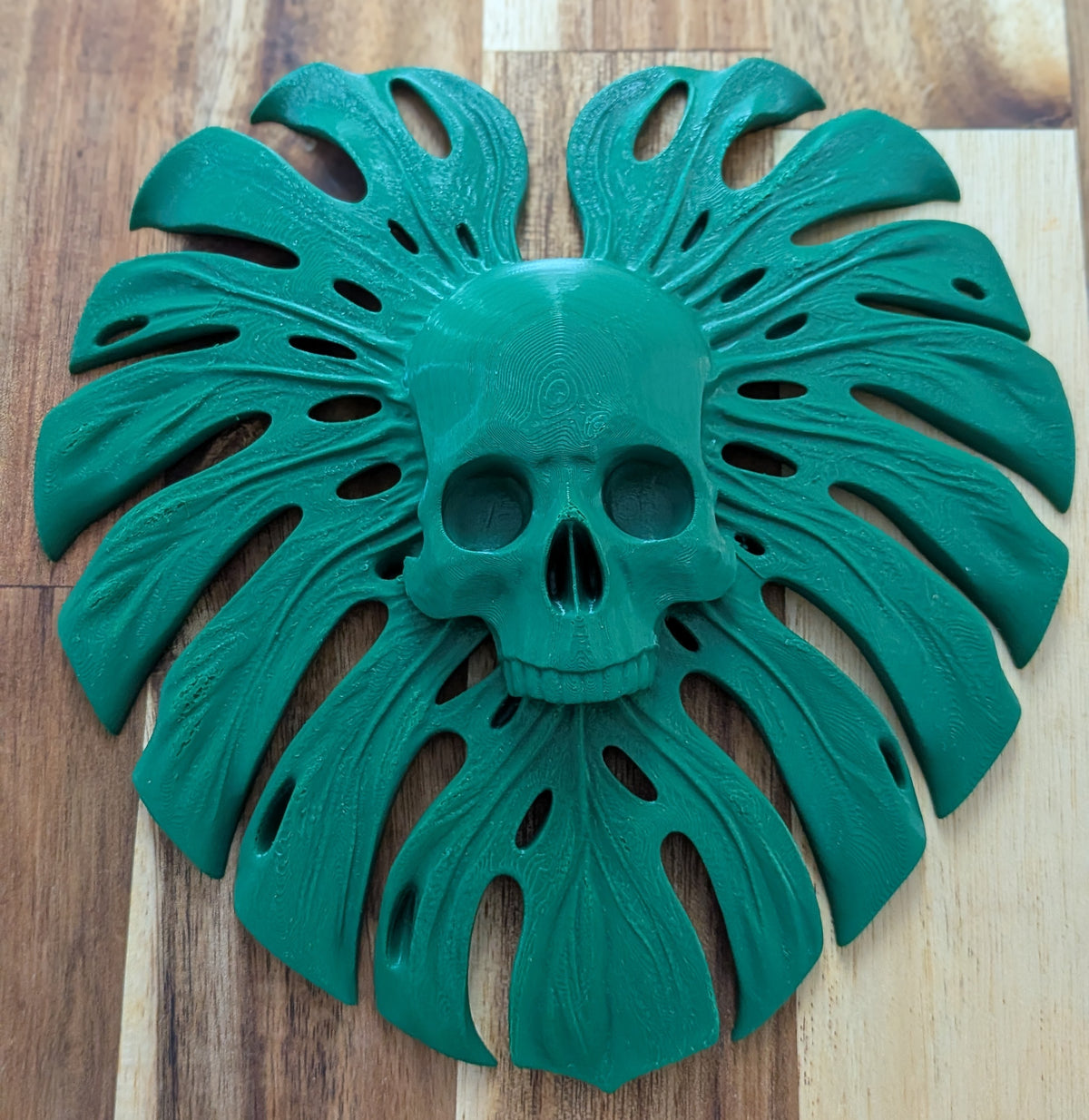 A 3D-printed wall art piece featuring a central skull surrounded by a monstera leaf design, in a vibrant teal green color, displayed on a wooden surface.