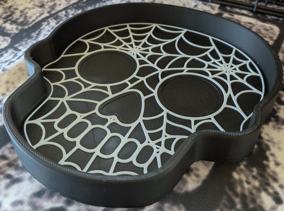 A black skull-shaped tray with a spiderweb and eye cutout design, photographed on a mottled beige and gray surface.