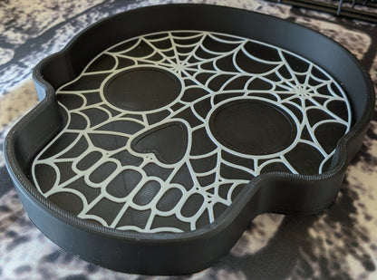 A black skull-shaped tray with a spiderweb and eye cutout design, photographed on a mottled beige and gray surface.