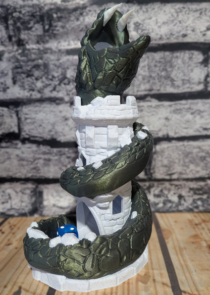 A fantasy sculpture of a dark green dragon coiled around a white tower, with a blue dice roller inside a ring at the base, set on a wooden surface against a gray brick wall background.