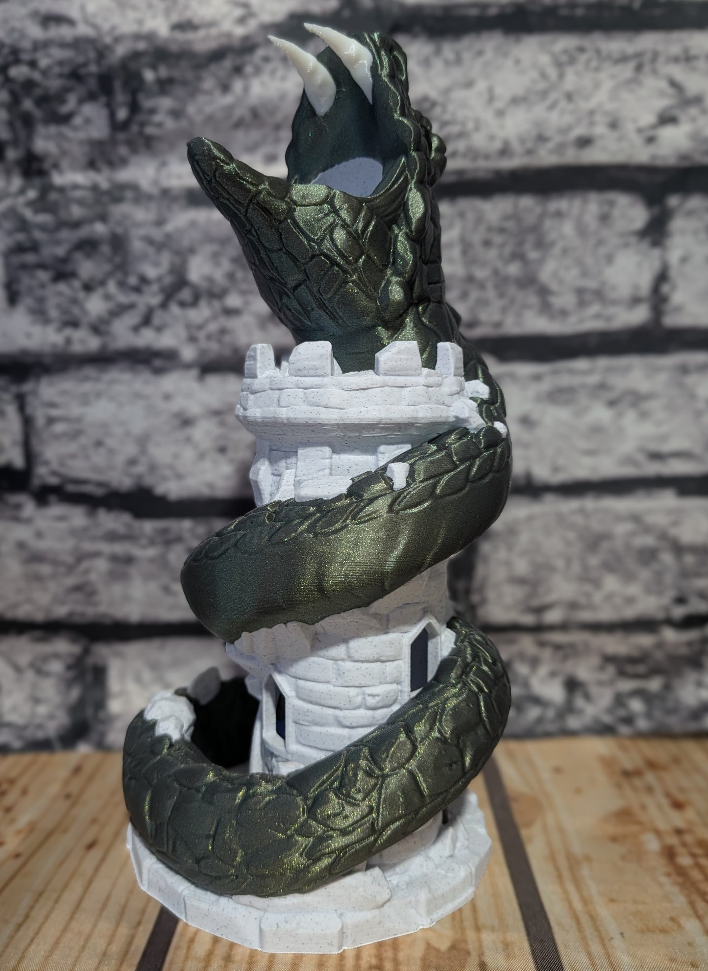 A dark green dragon figurine coiled around a white castle tower, displayed on a wooden surface against a gray brick wall background.