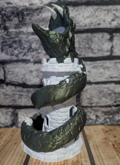 A dark green dragon figurine coiled around a white castle tower, displayed on a wooden surface against a gray brick wall background.