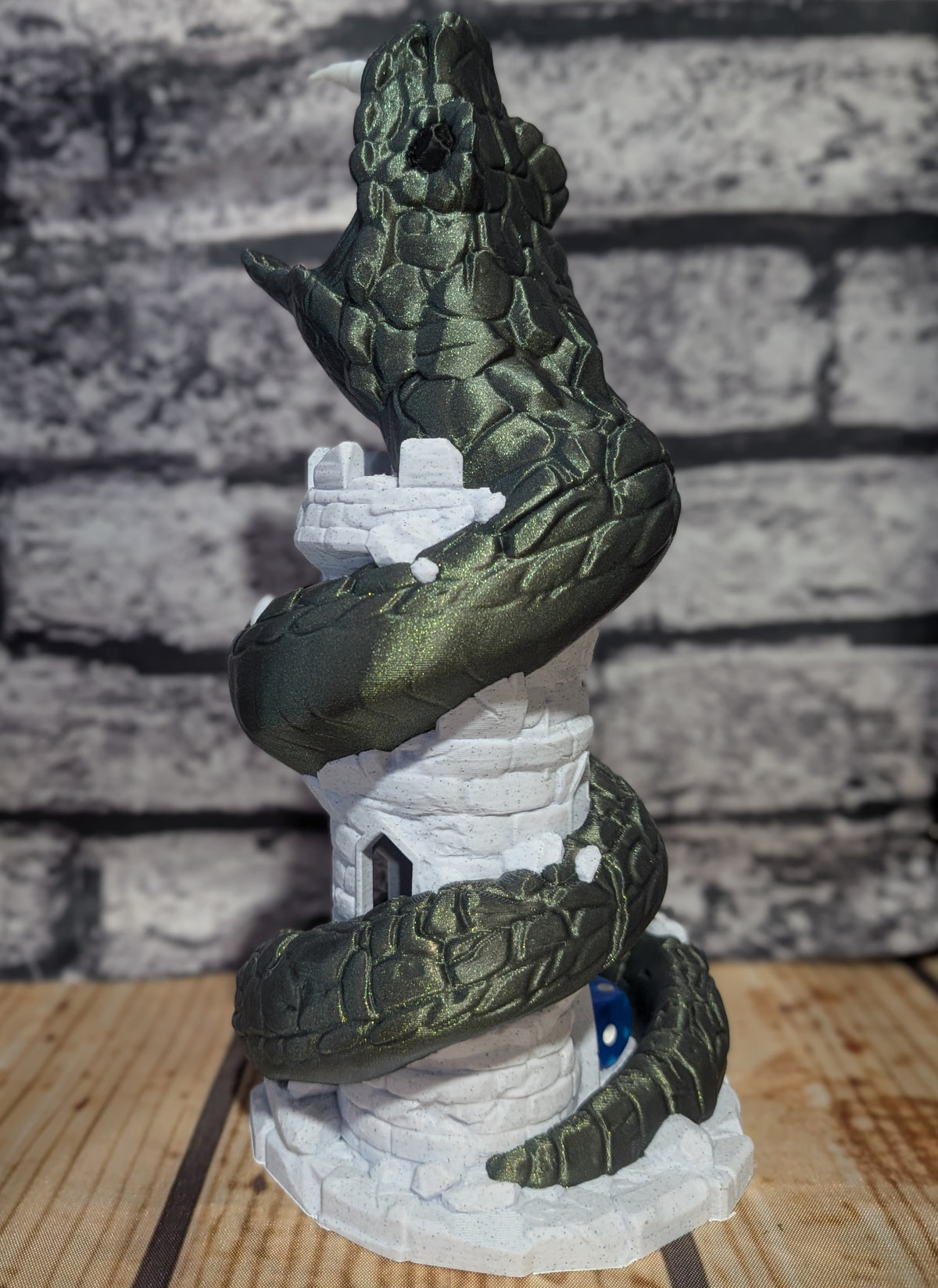A 3D-printed model of a green cobwebbed snake coiled around a white stone tower, displayed on a wooden surface against a gray brick wall background.