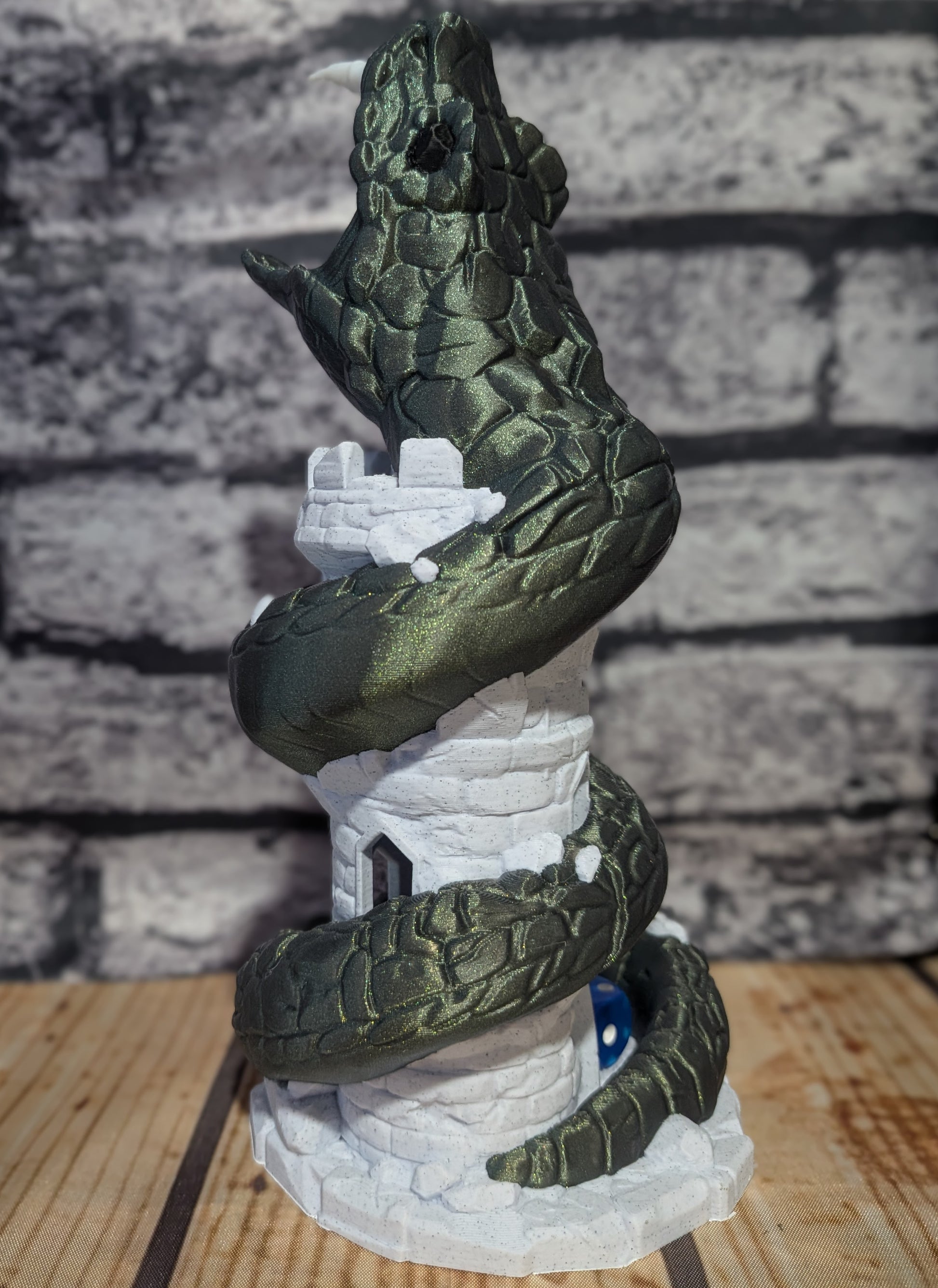 A 3D-printed model of a green cobwebbed snake coiled around a white stone tower, displayed on a wooden surface against a gray brick wall background.