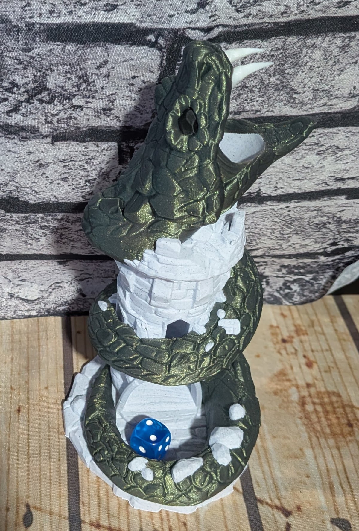 A 3D-printed fantasy sculpture of a green dragon coiled around a white stone tower, with a small blue die resting in its mouth, set against a faux-brick background.